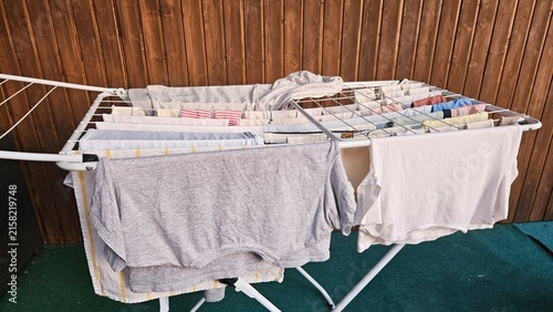 A white folding clothes drying rack holds grey and white garments; small items are clipped on the upper tier. The backdrop features vertical wooden panels and a dark green floor