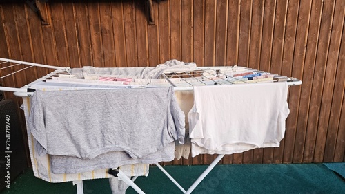 A white folding clothes drying rack holds grey and white garments; small items are clipped on the upper tier. The backdrop features vertical wooden panels and a dark green floor