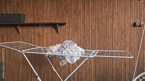 A white metal drying rack holds a crumpled grey garment and a small white cloth against warm wooden panels; a dark shelf and metal fittings accent the rustic setting