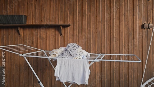 A white metal clothes drying rack holds light grey garments against a warm wooden wall; a dark shelf and rustic hardware accent the background