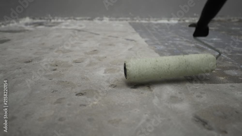 Low angle shot of paint roller moving across rough concrete floor applying primer. Action shows progression from start to finish with visible coverage change. Neutral gray tones dominate scene