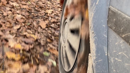 Low angle view of muddy car wheel spinning over leaf covered forest trail with blurred spokes, yellow leaf caught near tire, and scratched body panel with dried mud streaks