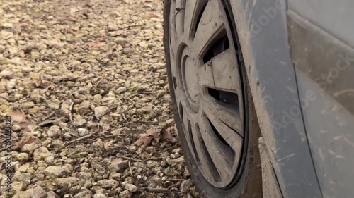 Dirty car wheel parked on rocky gravel, mud-splattered hubcap, compressed tire sidewall, and worn wheel arch shown from low angle with rough texture and overcast light