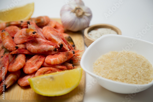 A close-up on a wooden board shows a handful of frozen shrimp with a slice of lemon, garlic, a salt shaker and a white bowl of rice are visible on a blurred background. Concept: highlighting seafood