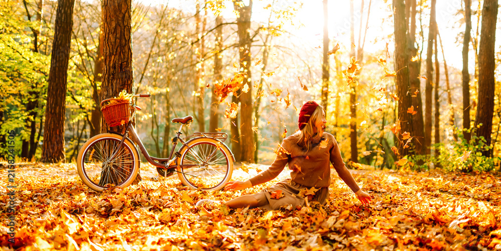 Fototapeta premium Woman is having fun in an autumn park, overlooking bicycle at sunset. Young woman throws bright yellow leaves while walking through sunny park. Concepts of relaxation, walking, and seasonality.
