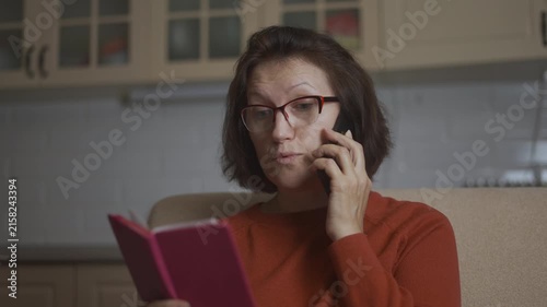 A woman sits on a sofa, holding a notebook while talking on a telefon. The woman appears focused on her telefon, glancing at the notebook for reference.