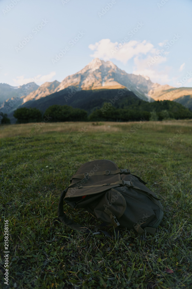Fototapeta premium Backpack resting on a sunlit field with distant mountain range, evoking outdoor adventure, hiking, and travel lifestyle in a calm nature scene with expansive landscape.