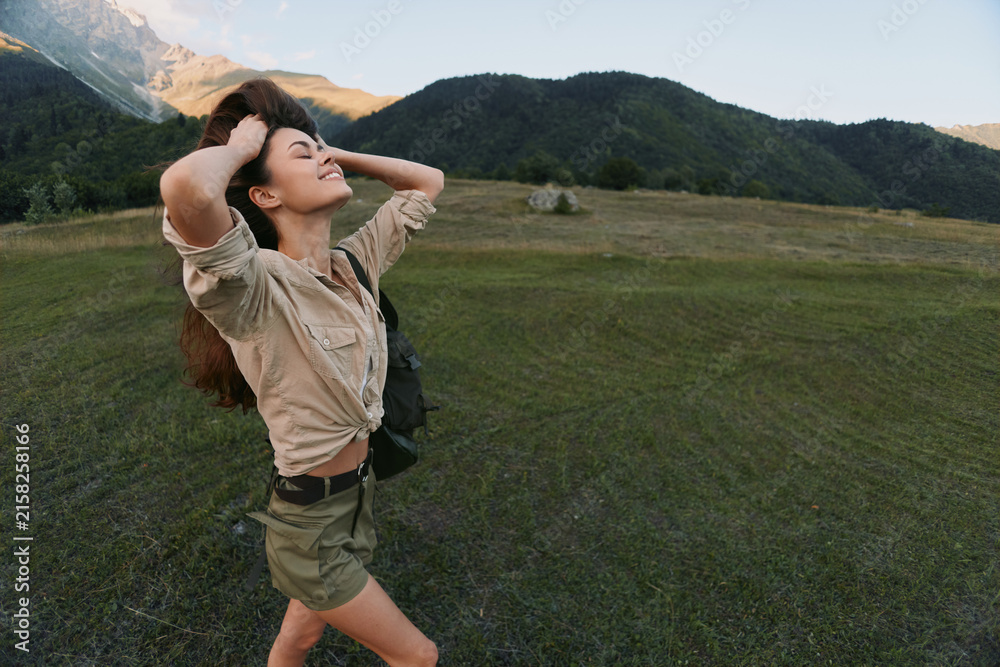 Fototapeta premium Woman enjoying outdoor life, standing in a meadow with arms raised, smiling, backpack visible. Nature mood, wellness, and freedom in a sunny landscape.