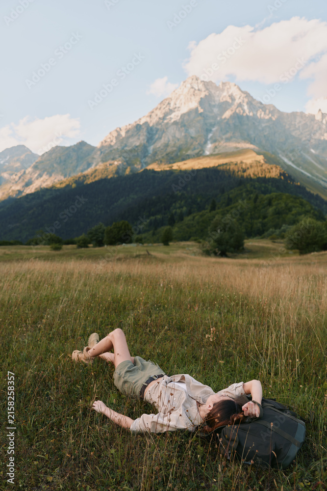 Fototapeta premium A woman rests in a sunlit meadow with a backpack beside her, mountains rising in the distance. A calm travel scene conveying rest, exploration, and connection with nature.