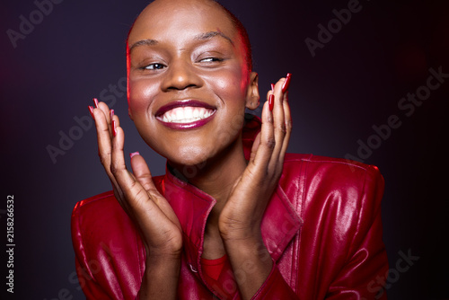 Foto Smiling African woman with dark skin and shaved head wearing a red leather jacket poses happily against a dark background