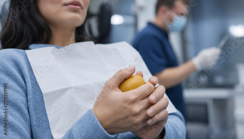 Medium close-up of an anxious female patient with fair skin holding a yellow stress ball tightly while sitting in a dental chair in a clinic with a blurred dentist in the background under soft indoor 