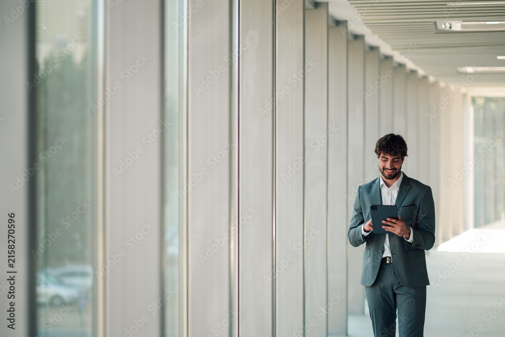 Fototapeta premium Professional businessman walking in modern office hallway, smiling and using digital tablet for work communication