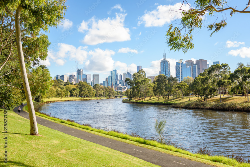 Naklejka premium Paved path on the bank of Yarra river on a sunny autumn day. Melbourne city centre skyline is visible in distance. 