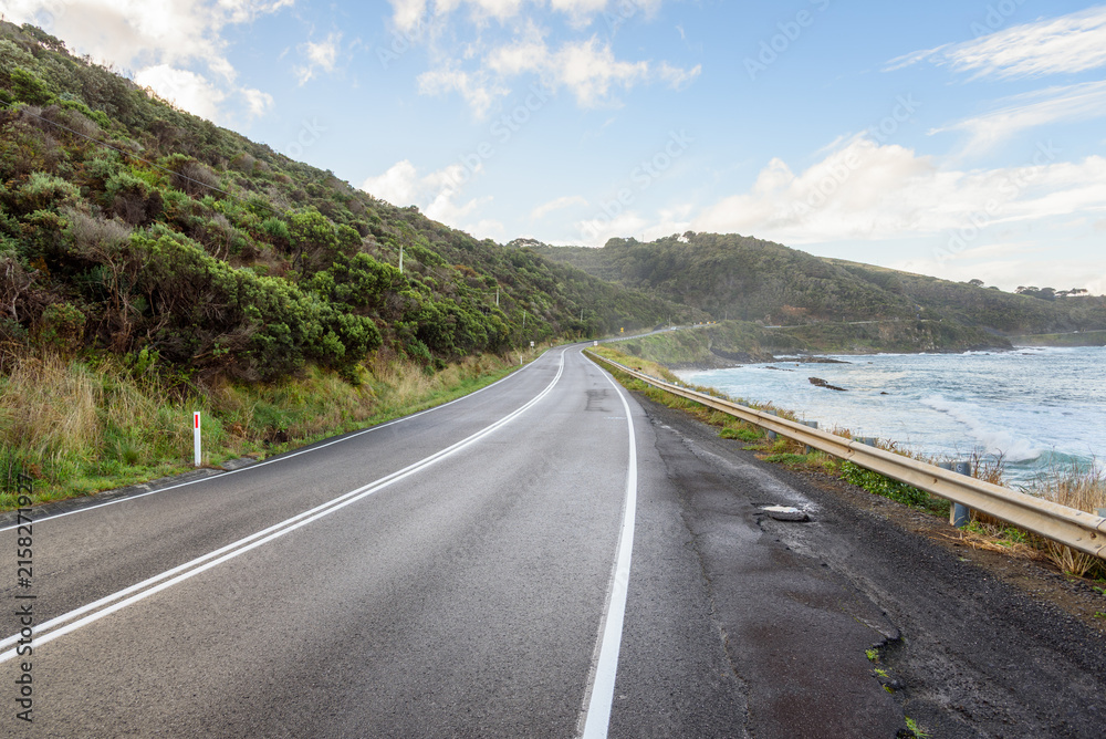 Fototapeta premium Deserted stretch of a coast road on a clear autumn day. A thin mist coming form the ocean hang over the road. Great Ocean Road, VIC, Australia.