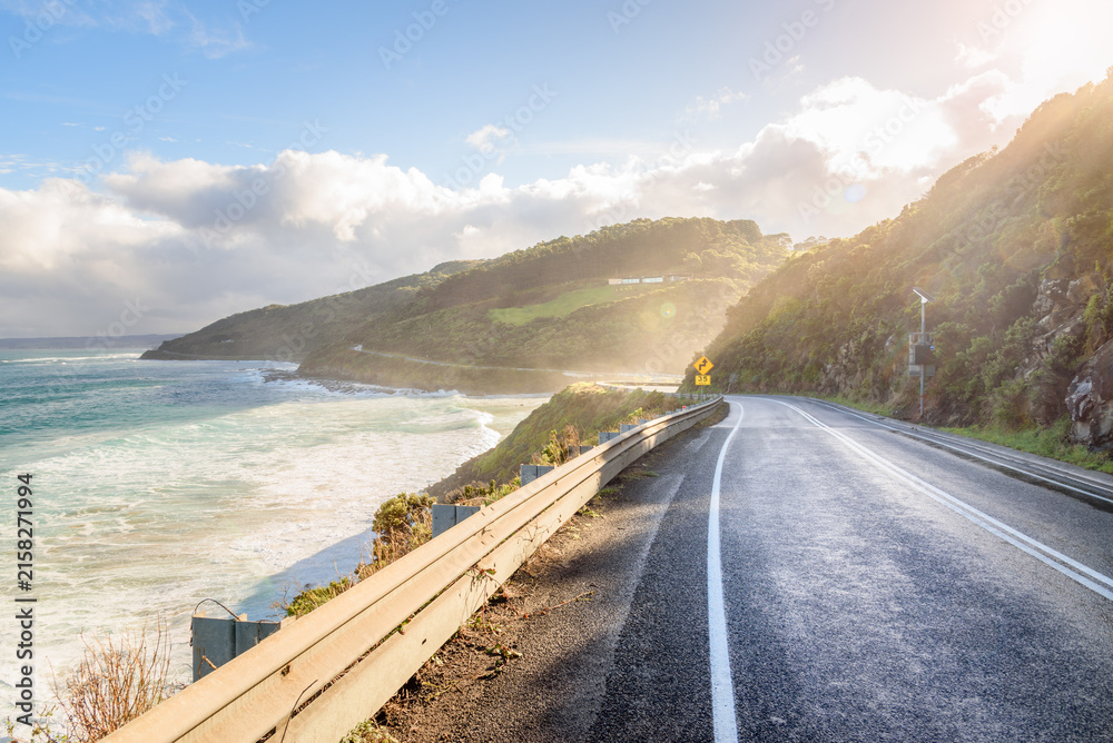 Fototapeta premium Empty stretch of the Great Ocean Road on the south-eastern coast of Australia at sunset in autumn. Mist coming form the ocean hangs over the road. Lens flare. 