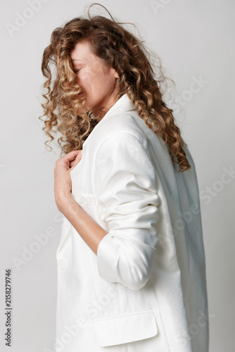 Authentic beauty of a middle aged woman with natural skin, curly hair, wearing a white blazer. Side profile on a light background. Calm, confident, unretouched.
