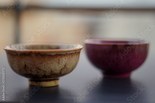 Two handcrafted ceramic tea bowls with earthy beige and deep burgundy glazes sit on a dark tabletop against a softly blurred background. The image represents contrast, companionship, balance, and the