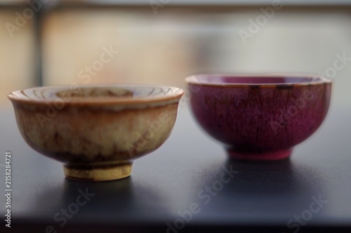 Two handcrafted ceramic tea bowls with earthy beige and burgundy glazes sit side by side on a dark tabletop. The image symbolizes companionship, contrast, balance, and the warmth of shared tea rituals