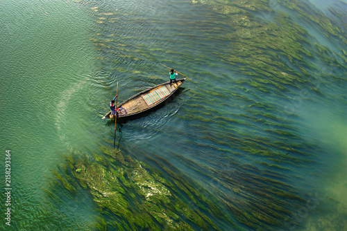 Aerial view of two fishermen in a traditional wooden boat navigating through lush green aquatic plants in the river in Sirajganj, Rajshahi Division, Bangladesh.