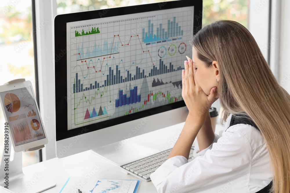 Fototapeta premium Stressed businesswoman covering face at desk with computer screen displaying financial graphs and data analysis