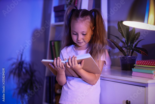 Young girl with pigtails reading a book in a dimly lit room, childhood educat...