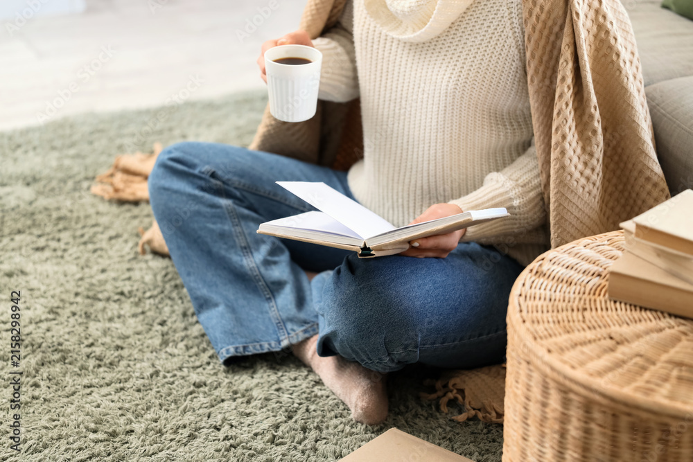 Obraz premium Young woman with book and cup of hot coffee sitting on carpet in living room