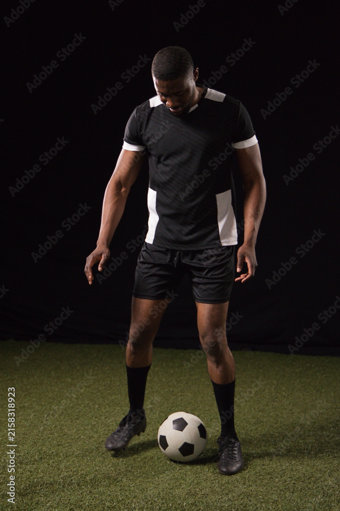 Fototapeta premium African American male standing on turf in studio wearing black-white soccer kit ball at feet