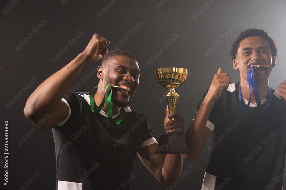 Fototapeta premium African male teammates celebrating in black-white jerseys, holding gold cup, biting medals in arena
