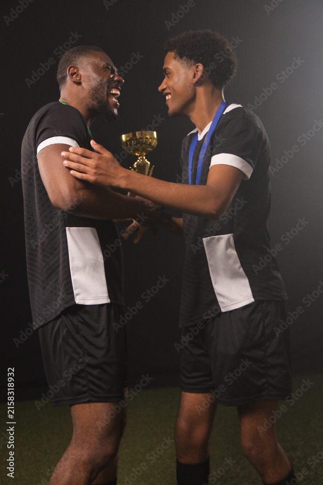 Fototapeta premium Male teammates wearing black-white jerseys holding gold trophy and blue ribbon on turf studio-lit