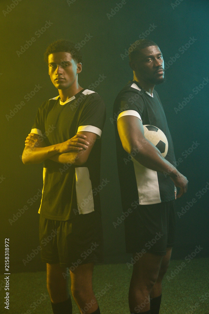 Fototapeta premium African male teammates posing back-to-back on turf, wearing black-white kits, holding soccer ball