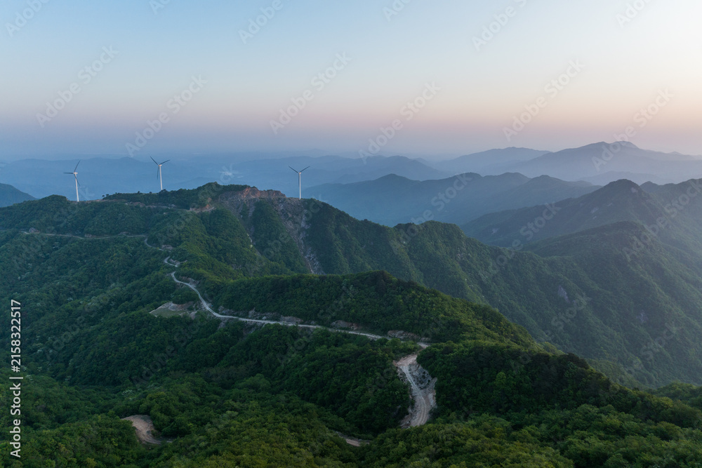 Fototapeta premium Layered green mountain ridges with wind turbines and a winding road, at dawn under a pale sky.