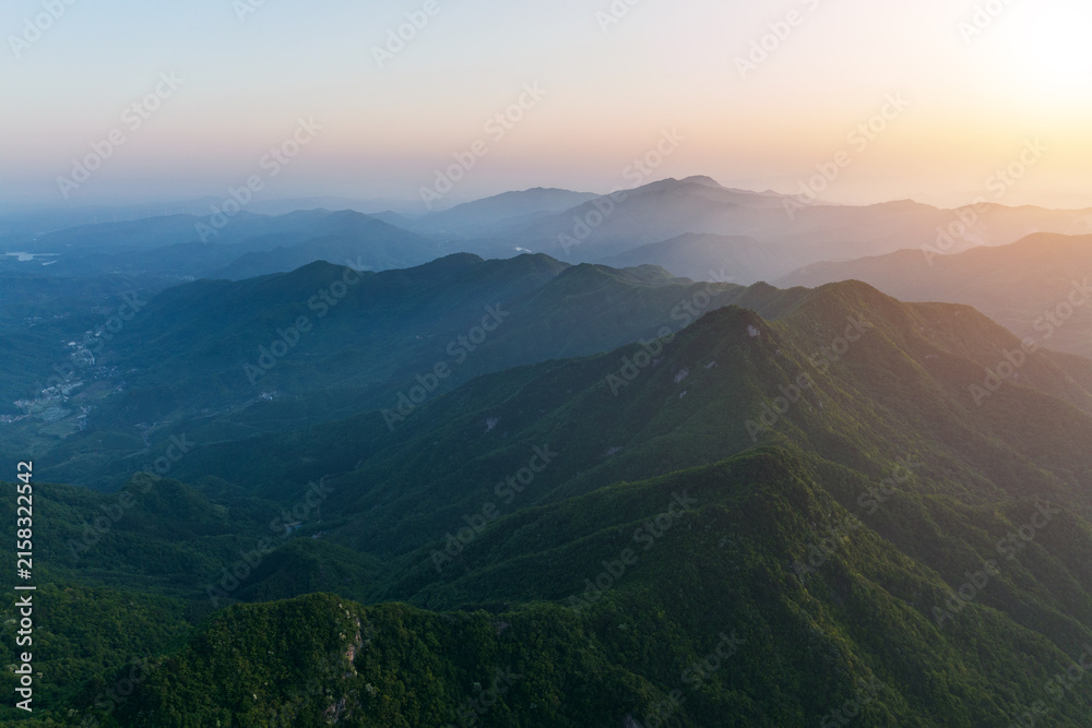 Fototapeta premium Layered forested mountain ranges stretch under a hazy sunrise sky, showing depth and natural landscape.