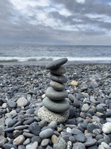 A stacked stone cairn balanced on a pebble beach against a blurred ocean and overcast sky background. Minimalist zen concept representing balance, harmony, and tranquility.
