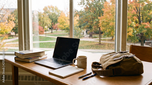 Window-side study desk with laptop, books, coffee mug and backpack overlooking autumn campus is perfect for back-to-school lifestyle backgrounds