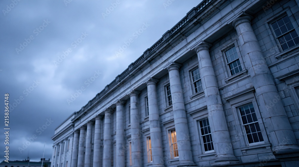 Fototapeta premium Low angle view of neoclassical building facade with tall columns and lit windows under dramatic cloudy evening sky