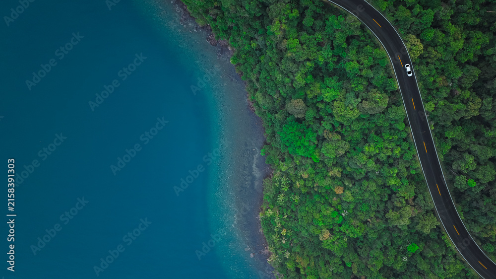 Fototapeta premium Top view aerial shot of a white car driving on a coastal road between lush green forest and turquoise ocean water, concept of scenic nature landscape representing travel and exploration. 