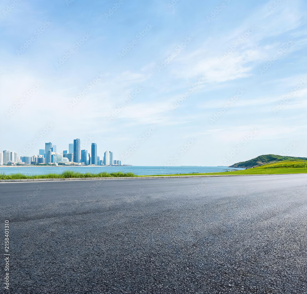 Fototapeta premium An empty asphalt road extends toward a coastal cityscape, with blue sea, green hill and clear sky in the view.
