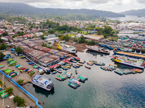 Panoramic aerial perspective of Bitung port in North Sulawesi. The lively harbor teems with passenger boats and ferries, seamlessly blending the urban coastal landscape with lush distant mountains.