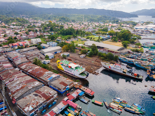 Panoramic aerial perspective of Bitung port in North Sulawesi. The lively harbor teems with passenger boats and ferries, seamlessly blending the urban coastal landscape with lush distant mountains.