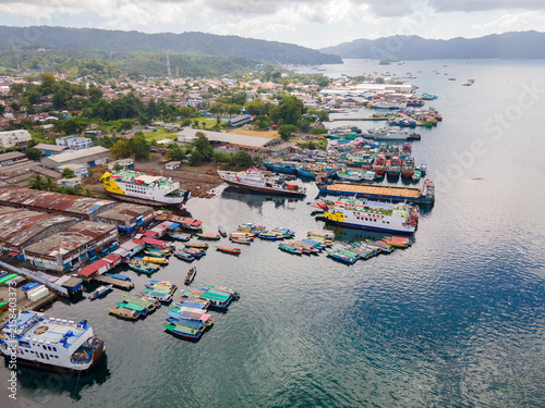 Panoramic aerial perspective of Bitung port in North Sulawesi. The lively harbor teems with passenger boats and ferries, seamlessly blending the urban coastal landscape with lush distant mountains.