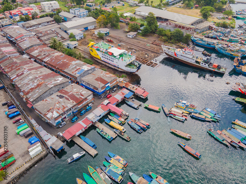 Panoramic aerial perspective of Bitung port in North Sulawesi. The lively harbor teems with passenger boats and ferries, seamlessly blending the urban coastal landscape with lush distant mountains.