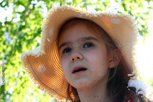 A young girl with light brown hair wearing a straw hat. She has a curious expression and is surrounded by greenery in a bright outdoor setting.