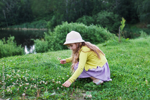 A young girl with long brown hair, wearing a yellow sweater and a pink hat, crouches on green grass, picking flowers near a calm lake surrounded by trees.