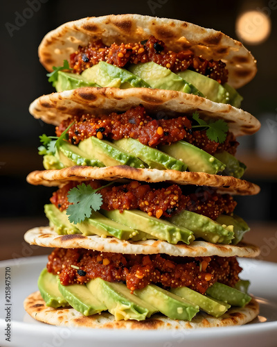 Harissa dip presented as a dramatic vertical tower, layers of crispy lavash alternating with thick harissa and avocado cream, tiny coriander leaves between layers, extreme close-up focusing
