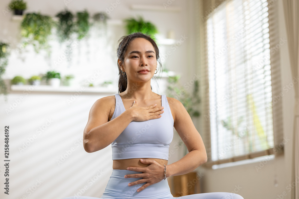Fototapeta premium Woman yoga meditation breathing home wellness exercise calm indoor plant Asian woman practicing yoga breathing exercise home with calm expression bright room with plant decor and window light