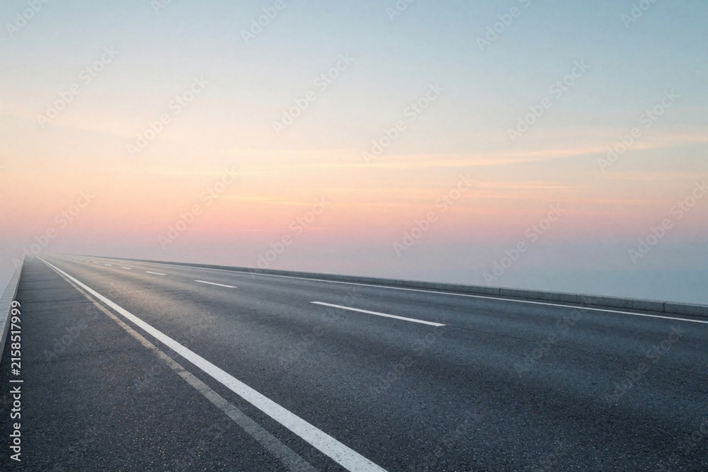 Fototapeta premium Empty asphalt highway stretches towards horizon under pastel sunset sky. Long straight road leads into distance with soft pink, blue colors. Minimalist landscape shows endless path for travel,