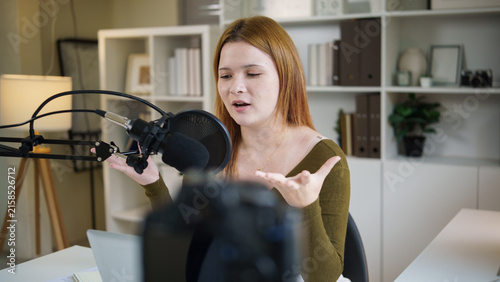 Bild auf Leinwand Young female podcaster speaking into a microphone while recording at home