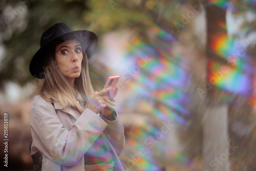 Surprised Young Woman Wearing Fedora Hat Holding Her Pink Smartphone. Shocked girl looks away with wide eyes while checking mobile device