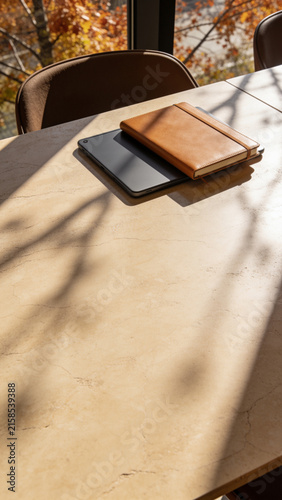 Brown leather notebook and digital tablet on marble table near window with autumn shadows in warm sunlight