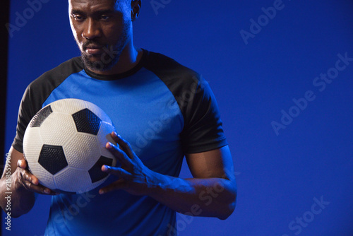 Mid-adult African male holding black-and-white soccer ball, posing in studio wearing blue shirt